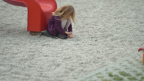 Ein-Kleines-Blondes-Mädchen-Sitzt-Neben-Der-Rutsche-Auf-Dem-Spielplatz-Im-Freien-Und-Spielt-Mit-Steinen