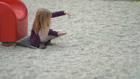 A-young-fair-haired-girl-sits-alone-near-the-slide-on-the-outdoor-playground-and-plays-with-stones