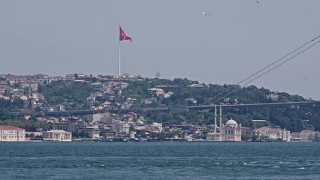 Turkish-flag-on-hillside-waving-in-breeze,-view-over-Bosphorus-strait-and-bridge