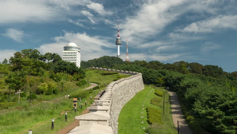 Seoul-City-Wall-In-Namsan-Park-and-N-Tower-Against-Blue-Sky-with-Floating-Clouds,-Korean-People-Walking-Up-and-Down-on-Mountain-Trails-on-Summer-Day