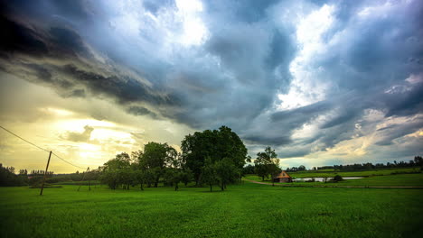 small-farm-in-meadow,-moving-clouds-timelapse,-moody-sky,-yellow-sunset,-big-green-tree-in-grassland,-small-lake-time-lapse,-establishment-shot,-rural-area,-countryside-farmland