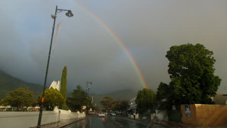 View-through-windscreen-as-car-drives-through-heart-of-Franschhoek-on-rainy-day-with-double-rainbow-in-sky