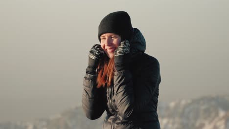 Young-caucasian-woman-in-beanie-adjusting-her-winter-jacket-during-sunrise-on-top-of-a-mountain