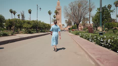 Young-caucasian-woman-in-a-dress-walking-towards-Koutoubia-Mosque,-Marrakech,-Morocco