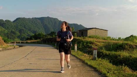 Frontal-tracking-view-of-asian-woman-carrying-hiking-backpack-across-countryside-pathway-at-sunset