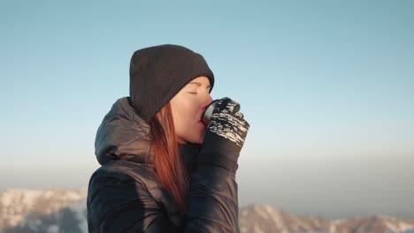Young-caucasian-woman-with-a-winter-jacket-and-beanie-sipping-tea-out-of-a-thermoflask-in-the-mountains