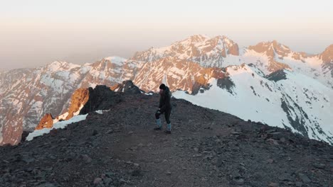 Young-caucasian-woman-in-winter-clothes-walking-up-a-mountain-during-sunrise-in-High-Atlas,-Mount-Toubkal,-Morocco