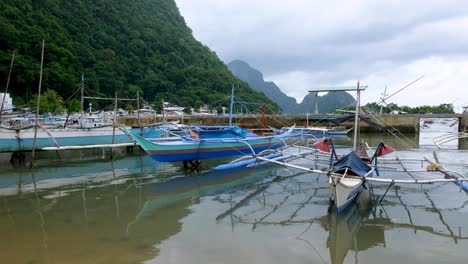 Traditional-outrigger-fishing-and-tour-boats-in-harbour-against-steep-cliffs-of-tropical-island-in-El-Nido,-Palawan,-Philippines