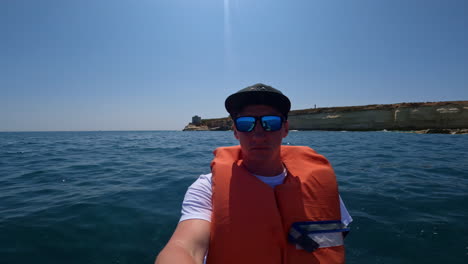 Junger-Mann-Im-Selbstporträt-Mit-Schwimmweste-Auf-Einer-Bootstour-In-Malta-Mit-Meer-Und-Klippen-Im-Hintergrund