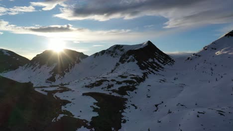 Sun-rays-breaks-out-over-snowy-mountain-in-scenic-winter-alpine-landscape,-drone