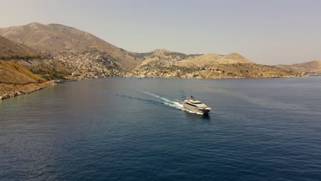 Luxurious-yacht-leaves-the-city-Symi-in-Greece-during-sunset,-aerial-pan-right