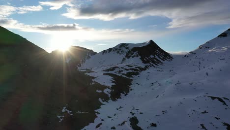 Drone-riser-reveals-sunrise-sun-rays-bursting-over-mountains,-snowy-Italian-Alps