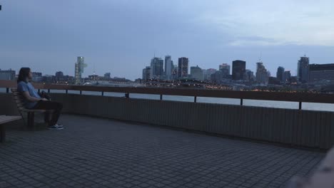 Man-with-Long-Hair-Sitting-on-a-Bench-Looking-Across-the-River-at-the-City-Skyline-in-Montreal,-Quebec,-on-a-Cloudy-Evening