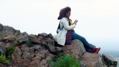 mixed-race-woman-with-curly-hair-is-sitting-on-the-ledge-of-a-cliff-in-Iceland-during-an-overcast-day