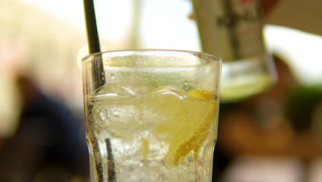 Pouring-carbonated-water-from-a-bottle-into-a-transparent-glass-with-ice,-lemon,-and-a-black-paper-straw
