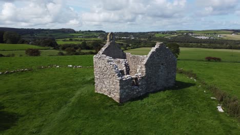 Establishing-aerial-view-Capel-Lligwy-ruined-chapel-on-Anglesey-island-green-countryside-coastline,-North-Wales
