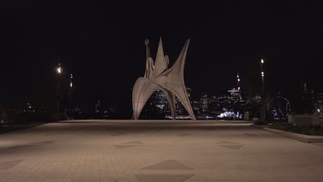 Art-Statue-at-Night-in-Parc-Jean-Drapeau-in-Montreal,-Quebec,-With-the-Illuminated-City-Skyline-in-the-Background