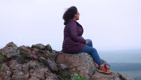 mixed-race-woman-with-curly-hair-is-sitting-on-the-ledge-of-a-cliff-in-Iceland-during-an-overcast-day