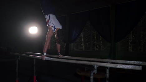 Professional-gymnast-in-a-dark-room-in-sportswear-stands-on-crayfish-on-parallel-bars-and-performs-flips-in-slow-motion.-Hard-training-in-preparation-for-the-Olympic-games