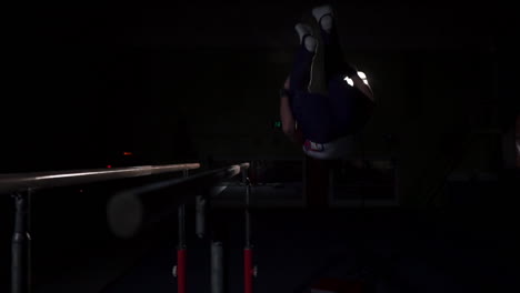Male-gymnast-acrobat-performs-handstand-on-parallel-bars-in-a-dark-room-in-slow-motion-sharing-a-somersault-and-landing-on-the-floor.-Training-before-the-Olympic-games.-A-professional-athlete