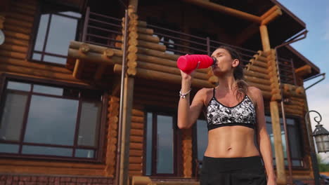 Sporty-brunette-woman-with-nice-abs-in-black-top-is-wet-from-sweat-on-the-background-of-wooden-mansion-after-a-workout,-drinks-water-from-a-bottle