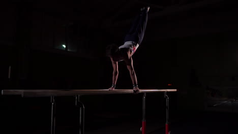 Professional-gymnast-in-a-dark-room-in-sportswear-stands-on-crayfish-on-parallel-bars-and-performs-flips-in-slow-motion.-Hard-training-in-preparation-for-the-Olympic-games
