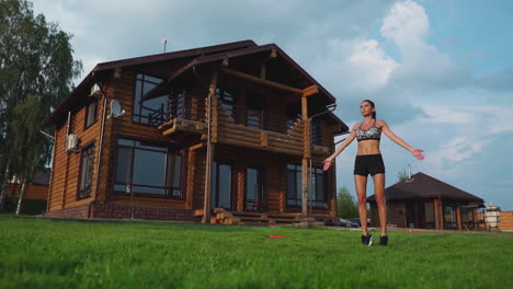 Slim-beautiful-woman-in-sportswear-on-the-background-of-the-house-on-the-lawn-performs-jumps-for-cardio-training-and-fat-burning