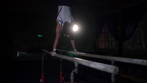 Male-gymnast-acrobat-performs-handstand-on-parallel-bars-in-a-dark-room-in-slow-motion-sharing-a-somersault-and-landing-on-the-floor.-Training-before-the-Olympic-games