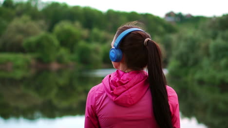A-girl-in-a-pink-jacket-and-blue-headphones-stands-with-her-back-and-looks-at-the-river-early-in-the-morning-after-a-run