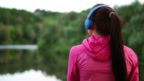 A-girl-in-a-pink-jacket-and-blue-headphones-stands-with-her-back-and-looks-at-the-river-early-in-the-morning-after-a-run