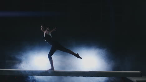 The-girl-performs-a-trick-on-a-log-in-backlight-and-slow-motion-in-sports-gymnastic-clothing.-Smoke-and-blue.-Jump-and-spin-on-the-balance-beam