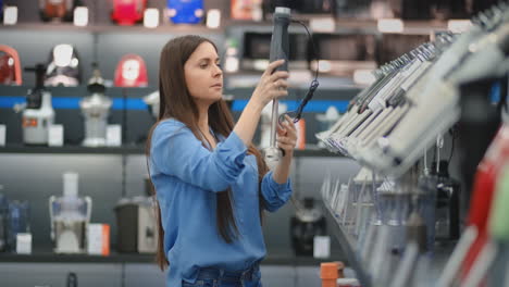 In-appliances-store-kitchen-appliances,-a-brunette-woman-in-a-blue-shirt-picks-a-blender-in-her-hands-and-considers-design-and-specifications.
