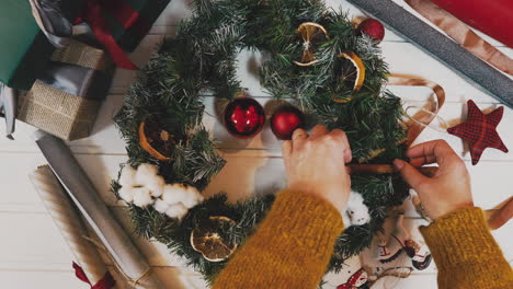 Draufsicht-Auf-Weibliche-Hände,-Die-Einen-Weihnachtskranz-Mit-Tannenzweigen-Und-Dekorativen-Beeren-Halten,-Mit-Dem-Wort-„Weihnachten“-Auf-Einer-Holztischplatte