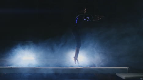 Professional-gymnast-girl-performs-a-jump-with-a-spin-on-the-balance-beam-in-slow-motion.-Smoke-blue-color-in-the-gym.-Gymnastics-competitions.