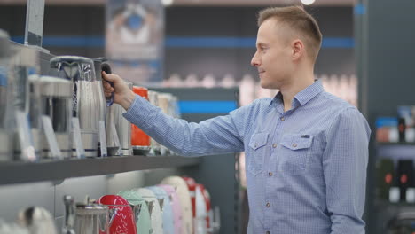 Young-handsome-man-chooses-an-electric-kettle-to-buy.-Inspects-the-device,-examines-the-price-tags-and-the-characteristics-of-the-model.