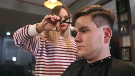 Interior-shot-of-working-process-in-modern-barbershop.-Side-view-portrait-of-attractive-young-man-getting-trendy-haircut.-Male-hairdresser-serving-client,-making-haircut-using-metal-scissors-and-comb.