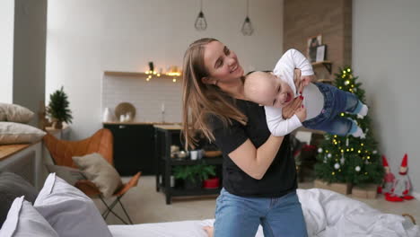 Lets-fly-Cheerful-beautiful-young-woman-holding-baby-girl-in-her-hands-and-looking-at-her-with-love-while-sitting-on-the-couch-at-home