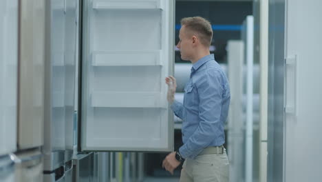 Der-Mann-öffnet-Die-Tür-Des-Kühlschranks-Im-Elektrofachmarkt-Und-Vergleicht-Ihn-Mit-Anderen-Modellen,-Um-Das-Neue-Haus-Zu-Kaufen