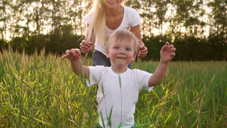 The-concept-of-a-happy-family.-In-the-rye-field-the-kid-walks-across-the-field-in-the-sun-setting-sun-looking-into-the-camera-Mom-stands-behind