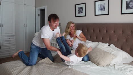 Mom-and-father-tickling-her-child.-people,-family,-fun-and-morning-concept---happy-child-with-parents-tickling-in-bed-at-home.-Happy-family-spending-time-together-in-bedtime-playing-and-hugging