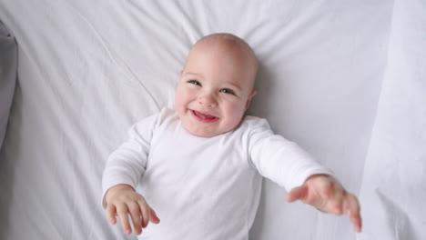 A-child-in-a-white-t-shirt-lying-on-a-white-bed-looking-at-the-camera-and-laughing-in-slow-motion,-top-view
