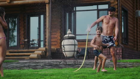 Mom-dad-and-two-sons-in-the-summer-on-the-grass-poured-water-from-a-hose-for-the-lawn.-Area-country-house-near-a-lake-at-sunset