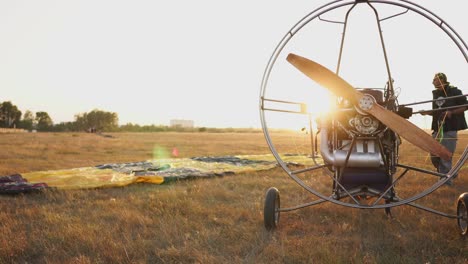 The-motor-paraglider-stands-in-the-field-at-sunset-with-a-wooden-propeller,-and-the-pilot-lays-out-the-parachute-and-aligns-the-slings