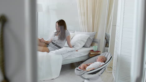 Mom-looks-into-the-mobile-phone,-and-next-to-the-baby-cradles-a-chair-in-a-white-interior.