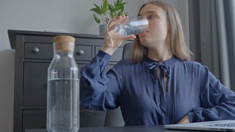 Woman-indoors-drinking-a-giant-glass-of-water-with-a-laptop-on-the-table