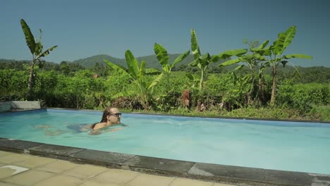 Caucasian-guy-swim-in-pool-surrounded-tropical-tree,-slowmo