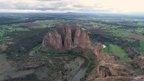 A-slow-moving-reverse-drone-shot-of-Smith-Rock,-Oregon,-a-river-that-flows-beside-it,-and-the-surrounding-farmland