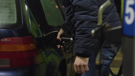 Man-pumps-gasoline-into-compact-dark-blue-car-on-cold-winter-night,-Petrol-station-foreground