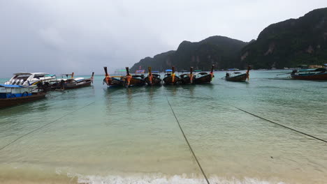 Traditional-longtail-boats-at-the-shore-of-the-Andaman-Sea-at-Phi-Phi-Island-in-rainy-weather,-Thailand,-Pullout-shot