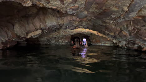 Girl-exploring-with-phone-flashlight-dark-caves-of-Roman-Baths-of-Casares-Manilva,-people-swimming-in-natural-spring-healing-sulfur-water-in-Spain,-good-health-benefits,-4K-shot